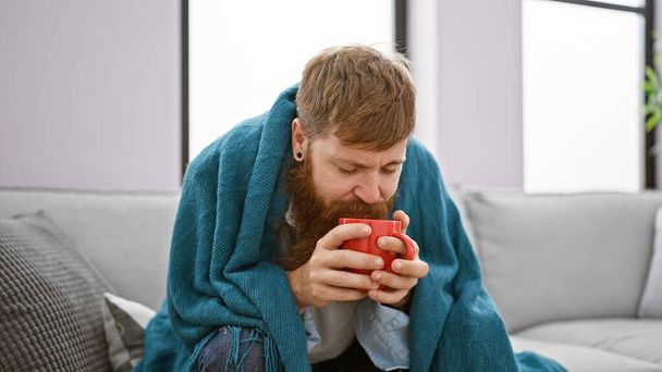 Csontig hűtött, fiatal, jóképű vöröshajú férfi forró kávét iszik, vigaszt és melegséget keres egy kényelmes takaró alatt a kanapén, megküzd a hideg téllel otthon. - Fotó, kép