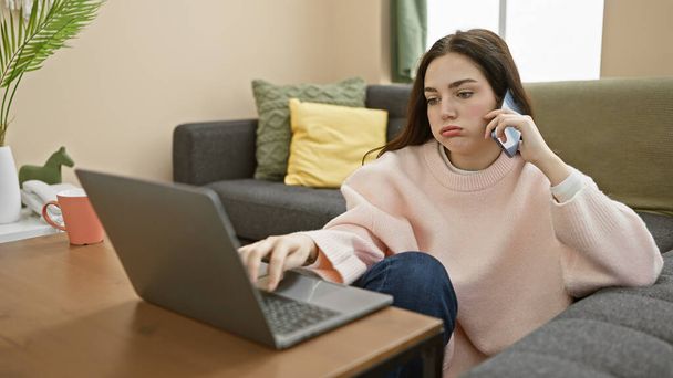 A young woman multitasking with a phone call and laptop in a cozy living room setting, appearing focused and casual. - Foto, Imagem
