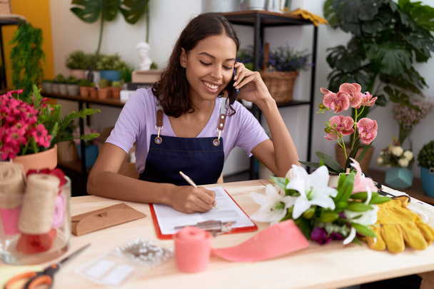 Young african american woman florist talking on smartphone writing on document at flower shop - Fotoğraf, Görsel