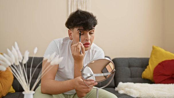 Young man applying makeup at home, reflecting on gender expression in a cozy, modern living room. - Photo, Image