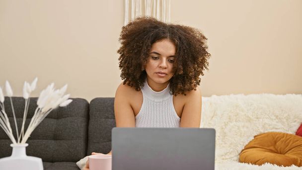 Ισπανίδα γυναίκα με σγουρά μαλλιά εργάζεται σε φορητό υπολογιστή στο άνετο σαλόνι - Φωτογραφία, εικόνα