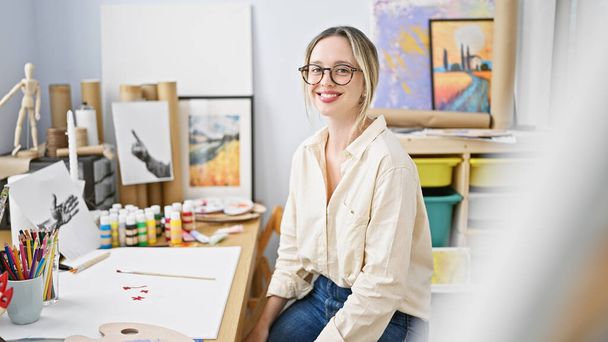 Jeune femme blonde artiste assise sur la table souriant au studio d'art - Photo, image
