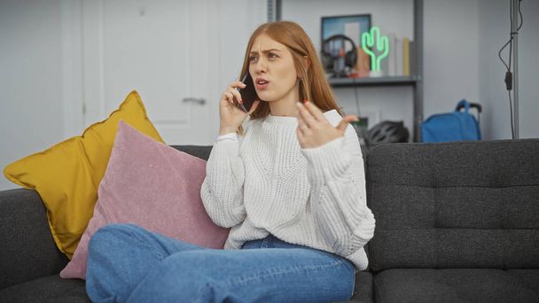 Zakłopotana młoda kobieta w białym swetrze i dżinsach rozmawia przez telefon siedząc na szarej kanapie w domu. - Zdjęcie, obraz