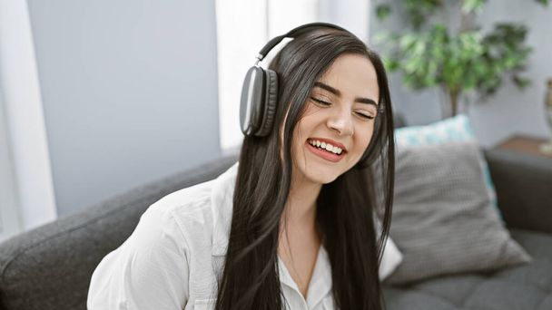 Fiatal spanyol nő élvezi a zenét fejhallgatóval egy hangulatos nappaliban, érzés örömteli. - Fotó, kép