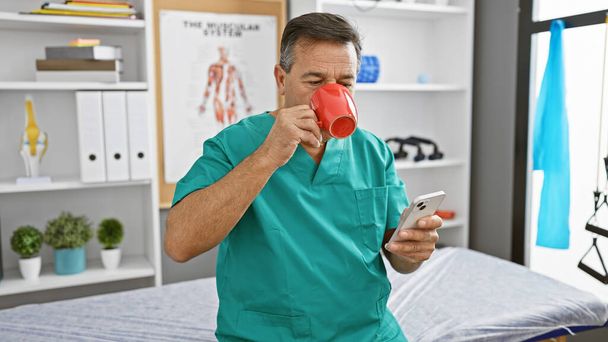 Volwassen man in scrubs het drinken van koffie en het controleren van de smartphone in een kliniek personeelskamer. - Foto, afbeelding