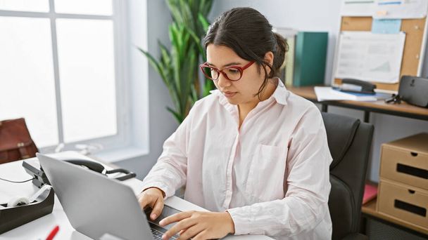 Hispanische Frau am Laptop in hellem, modernem Büro, mit Brille und weißem Hemd. - Foto, Bild