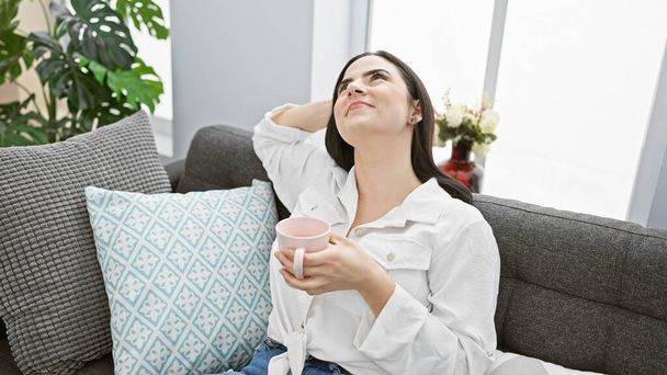 Uma jovem mulher hispânica relaxada desfruta de um momento de lazer com uma caneca em um ambiente acolhedor e moderno sala de estar. - Foto, Imagem