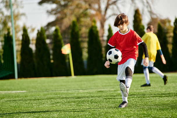 Młody chłopak w tętniącej życiem koszulce kopie piłkę nożną na zielonym polu pod jasnym słońcem. Jego skoncentrowana ekspresja pokazuje determinację i pasję do gry.. - Zdjęcie, obraz