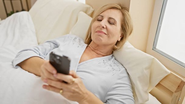 Eine ruhige Frau mittleren Alters entspannt mit dem Smartphone im Bett in einem gemütlichen Schlafzimmer und verkörpert Freizeit und moderne Konnektivität. - Foto, Bild