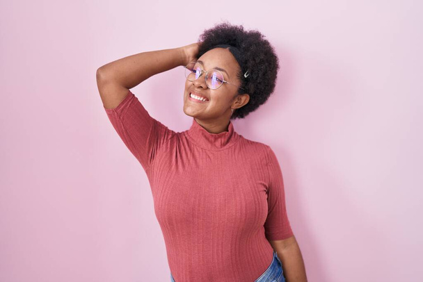Красивая африканская женщина с вьющимися волосами, стоящая на розовом фоне, улыбаясь уверенно трогательные волосы с жестом вверх рукой, позируя привлекательным и модным  - Фото, изображение