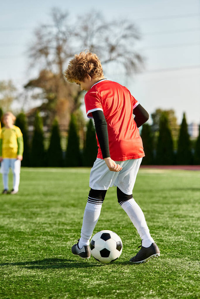 Egy fiatal fiú határozott arckifejezéssel rugdos egy focilabdát egy buja zöld mezőn a fényes nap alatt, kimutatva szenvedélyét a játék iránt.. - Fotó, kép