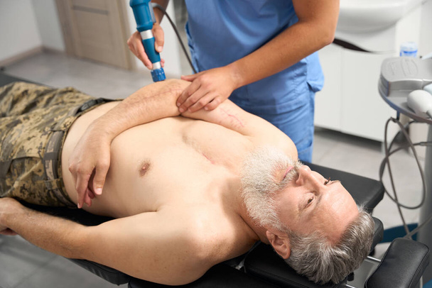Лікар, що працює з ліктем пацієнта з сірим волоссям, чоловік лежить на дивані з голим торсом - Фото, зображення