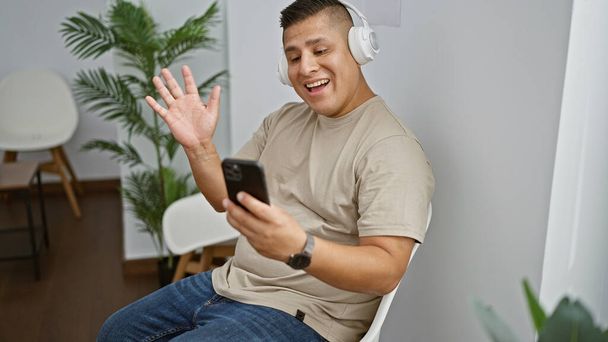 Allegro giovane latino gode di una videochiamata coinvolgente mentre è seduto in una sedia di attesa, abbracciando la comunicazione digitale - Foto, immagini