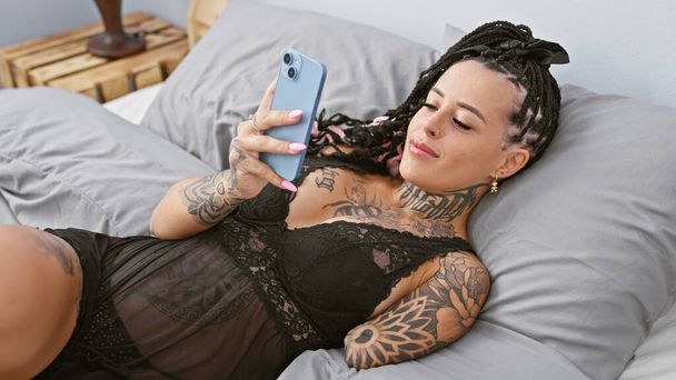 Mujer amputada hispana confiada enviando mensajes de texto en un teléfono inteligente, luciendo sexy en la luz de la mañana, acostada en el dormitorio - Foto, imagen