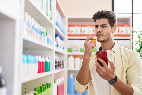 Молодий клієнт іспаномовного чоловіка за допомогою смартфона тримає пляшку таблеток в аптеці
 - Фото, зображення