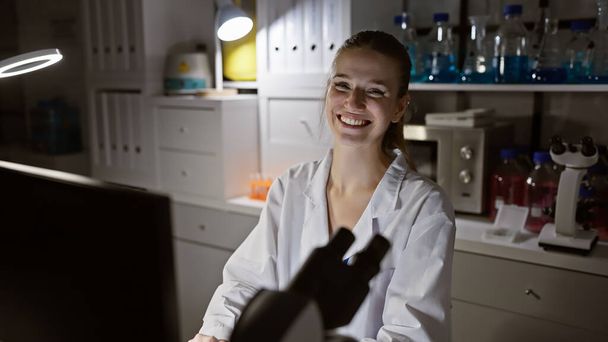 Χαμογελαστή νεαρή γυναίκα με εργαστηριακή ποδιά που εργάζεται σε σύγχρονο εργαστήριο με επιστημονικό εξοπλισμό - Φωτογραφία, εικόνα