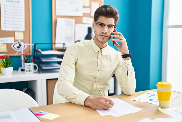 Young hispanic man business worker using smartphone writing on document at office - Photo, Image
