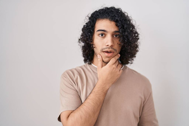 Hispanischer Mann mit lockigem Haar, der vor weißem Hintergrund steht und fasziniert vor Ungläubigkeit, Überraschung und staunendem Gesichtsausdruck mit den Händen am Kinn steht  - Foto, Bild