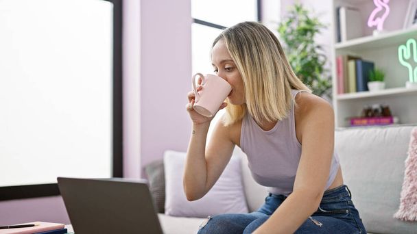 Laptop kullanan genç sarışın kadın evde kahve içiyor. - Fotoğraf, Görsel
