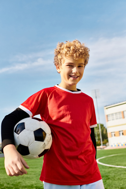 Młody chłopiec pewnie stoi na bujnym, zielonym boisku do piłki nożnej, trzymając piłkę z determinacją. Słońce świeci jasno, rzucając ciepły blask na jego gorliwą twarz. - Zdjęcie, obraz