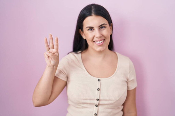 Fiatal spanyol nő áll a rózsaszín háttér mutatja, és rámutatva ujjak száma 3, miközben mosolyogva magabiztos és boldog.  - Fotó, kép