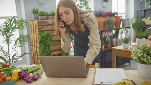 Eine junge Floristin in Schürze am Telefon, die ihr Geschäft mit einem Laptop inmitten frischer Blumen in einem Geschäft führt. - Foto, Bild