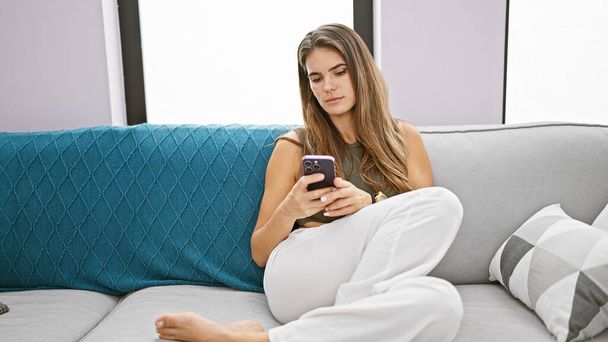 Młoda piękna Latynoska kobieta za pomocą smartfona siedzi na kanapie myśląc w domu - Zdjęcie, obraz