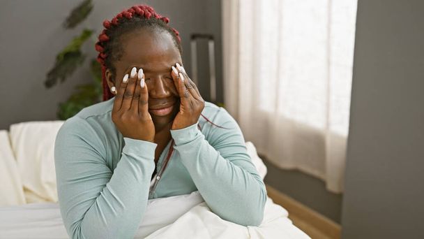 Взрослая черная женщина с косичками сидит на кровати в помещении, выражая головную боль или страдание в клинике - Фото, изображение