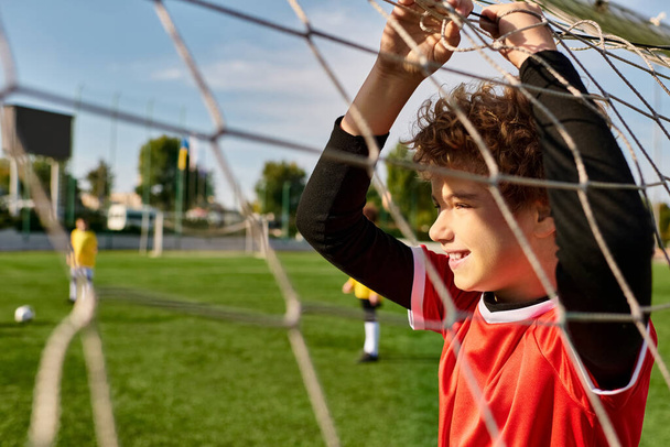 Ein kleiner Junge steht hinter einem Fußballnetz und hält einen Fußballball in den Händen. Sein fokussierter Blick deutet auf Entschlossenheit und Leidenschaft für den Sport hin, während er seine Torwartfähigkeiten übt. - Foto, Bild