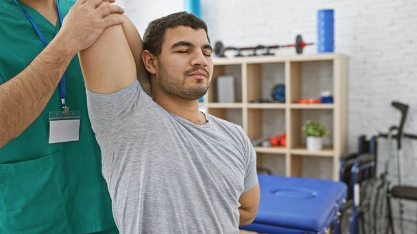 Un fisioterapista maschile in scrub assiste un uomo con esercizi di spalla in una clinica di riabilitazione illuminata. - Foto, immagini