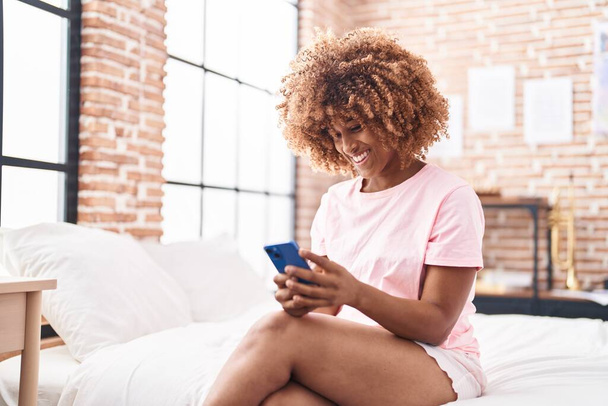 Mujer afroamericana usando teléfono inteligente sentado en la cama en el dormitorio - Foto, Imagen