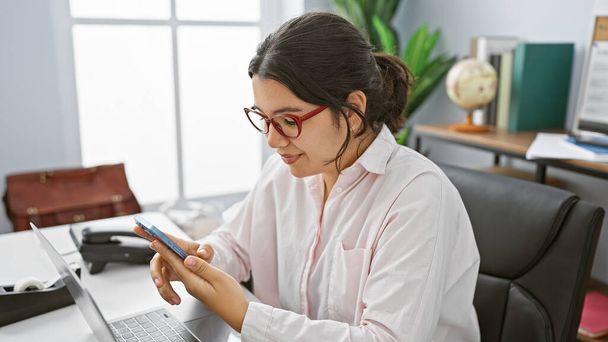 Uma mulher hispânica profissional focada em seu smartphone em um ambiente de escritório moderno. - Foto, Imagem