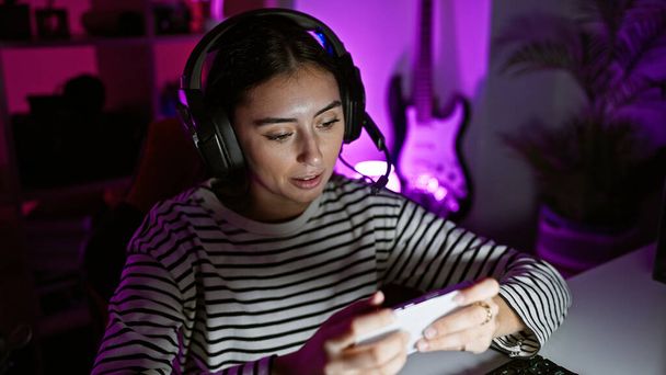 Una mujer hispana enfocada que usa auriculares y usa un teléfono inteligente en una sala de juegos iluminada por neón por la noche. - Foto, imagen