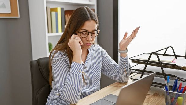 Una giovane donna con gli occhiali e una camicia a righe parla al telefono in un ambiente ufficio moderno, cercando preoccupato. - Foto, immagini