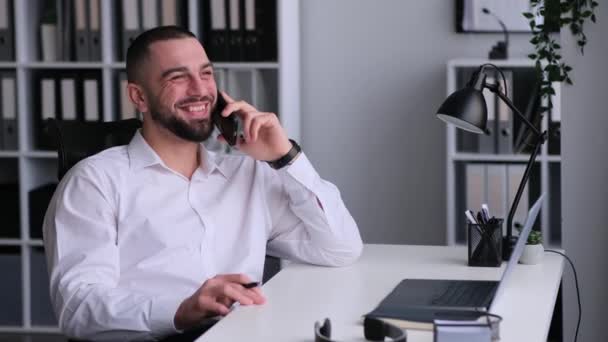Onnellinen valkoihoinen miesfreelancer puhuu puhelimessa ystävien kanssa, suunnittelee viikonloppua, nauraa ja rentoutua työpaikalla tauon aikana. - Materiaali, video