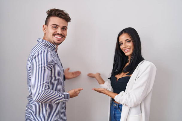 Jovem casal hispânico em pé sobre fundo branco convidando para entrar sorrindo natural com a mão aberta  - Foto, Imagem