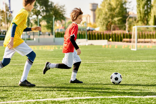 Twee jonge kinderen, die kleurrijke voetbaltruien dragen, voetballen enthousiast op een groen veld. Ze schoppen de bal heen en weer met precisie en enthousiasme, tonen hun passie voor de sport. - Foto, afbeelding