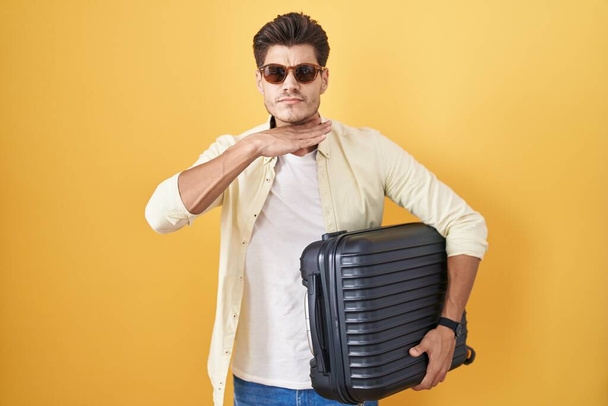 Jeune homme hispanique tenant une valise allant en vacances d'été tranchant la gorge avec la main comme un couteau, menace d'agression avec une violence furieuse  - Photo, image
