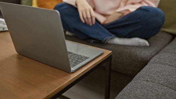 Egy fiatal nő véletlenül ül egy szürke kanapén egy nyitott laptop mellett egy hangulatos nappaliban.. - Fotó, kép
