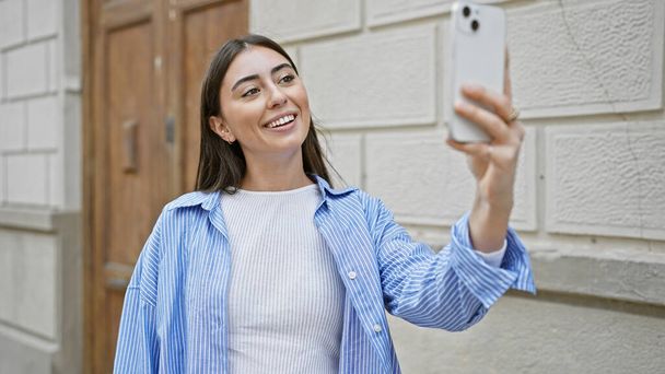 Eine junge hispanische Frau macht ein Selfie auf einer Straße in der Stadt und zeigt ihr attraktives Lächeln und ihren lässigen Stil. - Foto, Bild