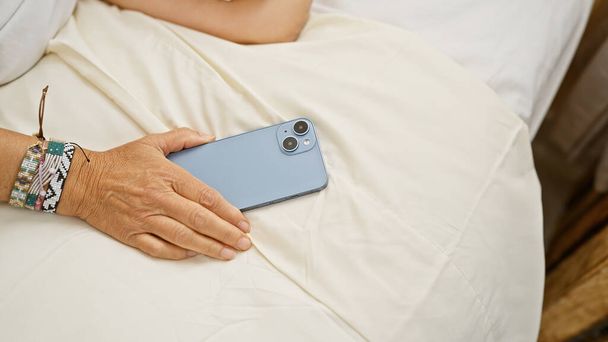 Крупним планом зріла жінка з браслетом тримає смартфон на ліжку в добре освітленій кімнаті, пропонуючи спокійну обстановку в приміщенні. - Фото, зображення