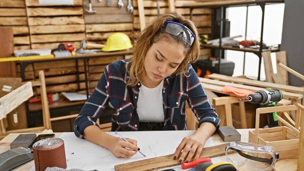 Atractiva joven carpintera hispana, vestida para mayor seguridad con gafas, anota hábilmente notas en su taller de carpintería - Foto, imagen