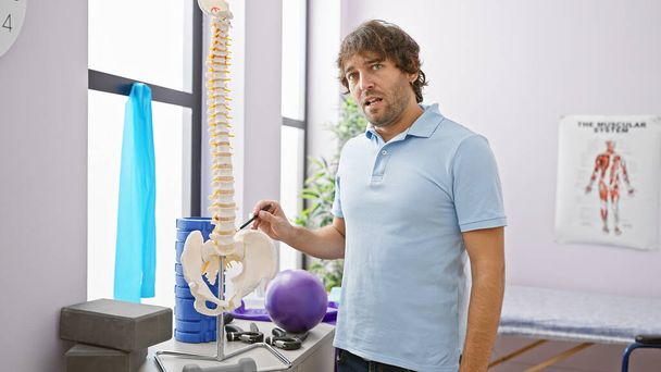 Hombre barbudo guapo apuntando al modelo de columna vertebral en una sala de rehabilitación clínica con equipo de ejercicio. - Foto, Imagen