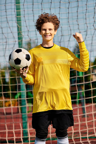 Ένα νεαρό αγόρι στέκεται μπροστά από ένα δίχτυ ποδοσφαίρου, κρατώντας μια μπάλα ποδοσφαίρου στα χέρια του, με ένα βλέμμα αποφασιστικότητας στο πρόσωπό του. - Φωτογραφία, εικόνα