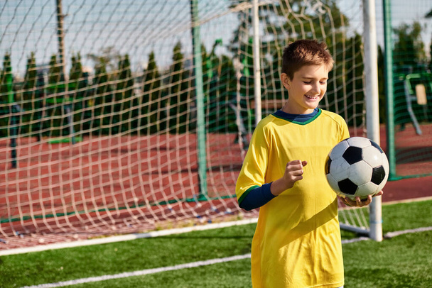 Ένα ταλαντούχο αγόρι φοράει μια φωτεινή κίτρινη στολή ποδοσφαίρου με αυτοπεποίθηση κατέχει μια μπάλα ποδοσφαίρου, αποπνέοντας πάθος και αποφασιστικότητα καθώς ετοιμάζεται για ένα παιχνίδι. - Φωτογραφία, εικόνα