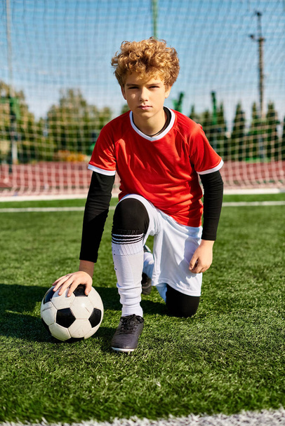 サッカーユニフォームの少年が優雅に草の上にひざまずき,彼の前でサッカーボールを保持する. - 写真・画像
