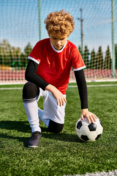 Um menino com cabelo escuro se ajoelha na grama, estendendo a mão para pegar uma bola de futebol. Seu foco é apenas na recuperação da bola, cercada pela vegetação do campo.. - Foto, Imagem