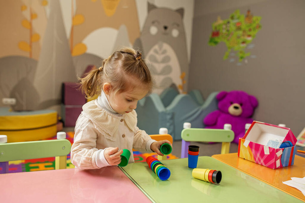 焦点を当てられた子供の少女は背景のおもちゃが付いている明るい遊び場のテーブルの多彩な積み重ねのコップと遊んでいます. - 写真・画像