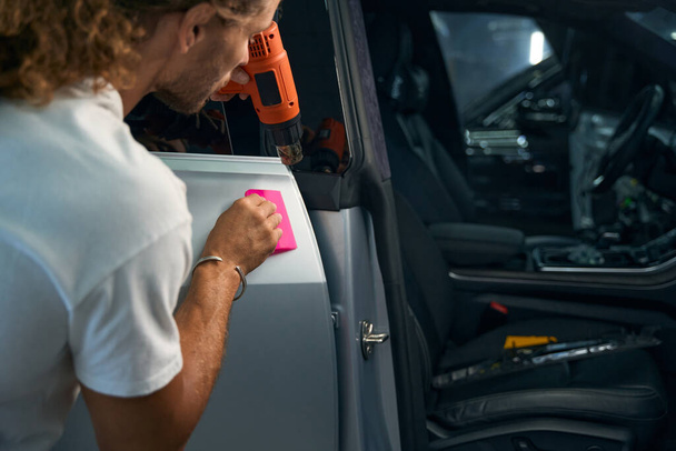 Maestro está en el proceso de aplicación de película anti-grava a las puertas del coche, utiliza una espátula rosa y aire caliente - Foto, Imagen