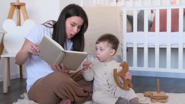 Blanke nieuwsgierige baby luistert naar liefhebbende moeder die hem een boek voorleest op de vloer in de speelkamer. Ontspannen en samen onderwijs geven. - Video